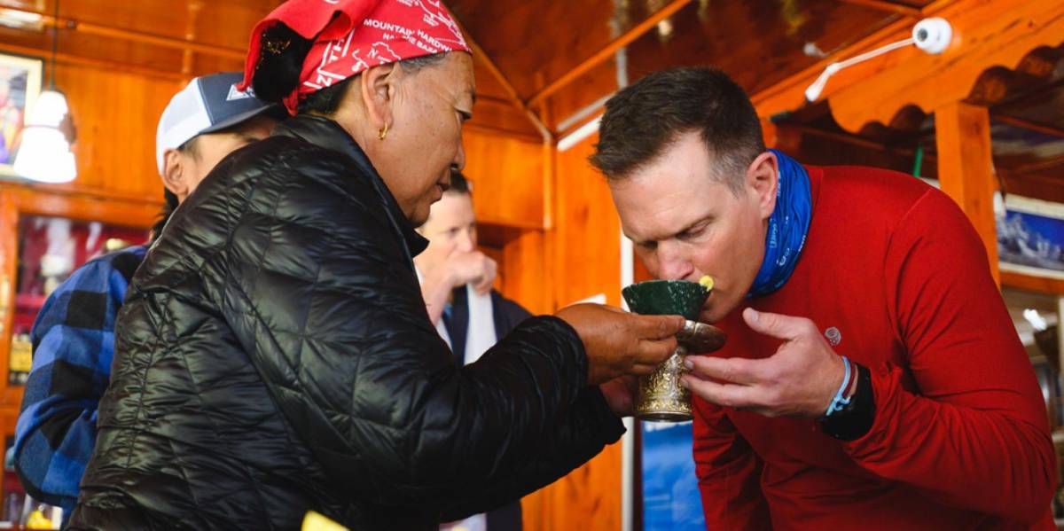 A woman in a black jacket and red scarf offers a decorative cup to a man in a red shirt, inside a cozy wooden setting.