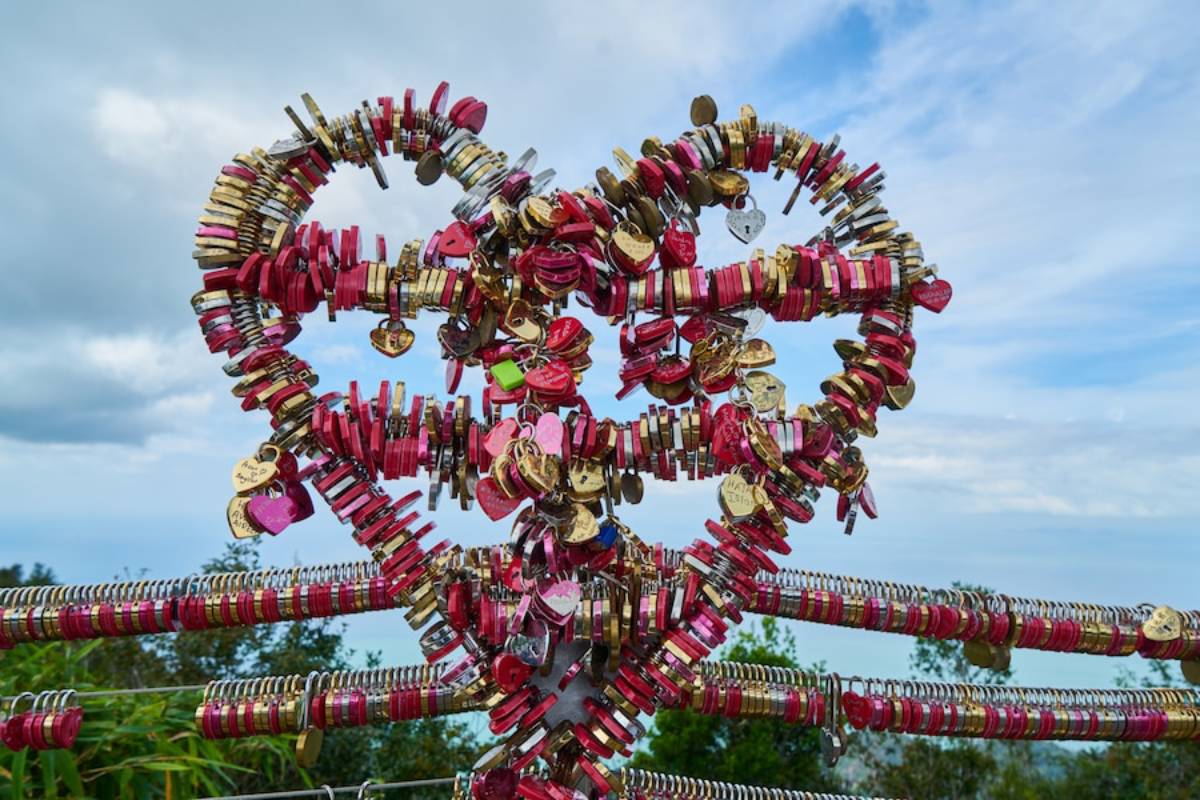A heart-shaped structure made of colorful love locks, symbolizing affection, with a scenic background of sky and greenery.