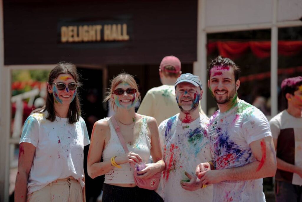 A group of four people, covered in vibrant colored powders, stand together in front of a sign that reads Delight Hall.