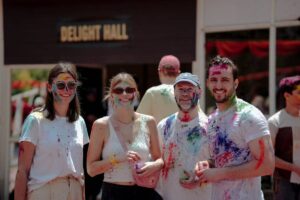 A group of four people, covered in vibrant colored powders, stand together in front of a sign that reads Delight Hall.