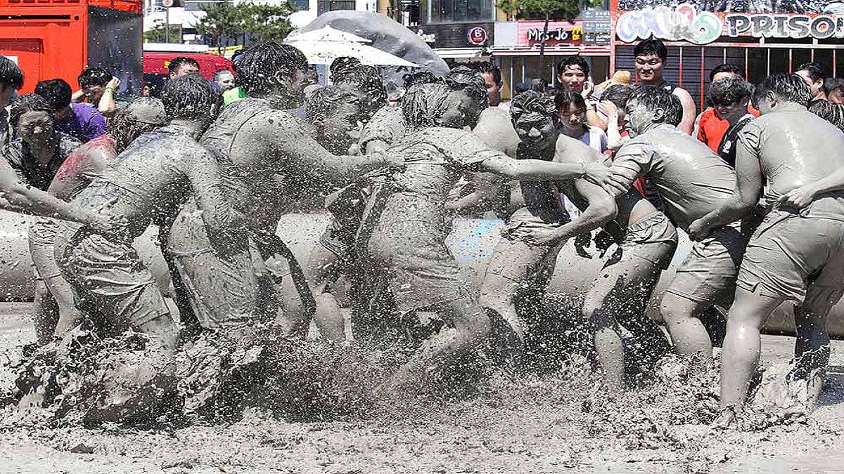 A group of muddy individuals engage in a playful tug-of-war on a sunny day, splashing water and mud around joyfully.