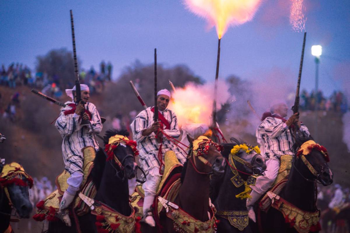 Four horsemen in traditional attire fire weapons into the air during a celebration, with a crowd watching in the background at dusk.