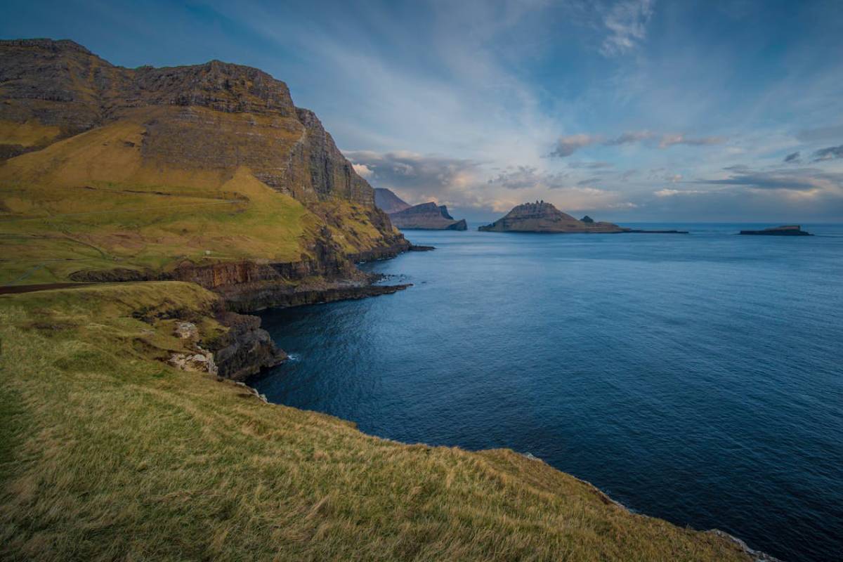 Coastal view of dramatic cliffs and serene waters, with distant islands under a blue sky and wispy clouds in the Faroe Islands.