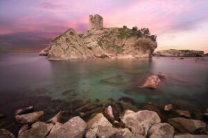 A serene coastal scene featuring a rocky island with a castle ruins, surrounded by calm turquoise waters and a vibrant sunset sky.