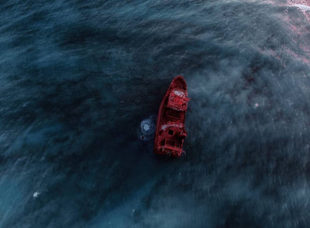 An aerial view of a rusted shipwreck adrift in dark blue waters, creating a stark contrast with its vibrant red hull.