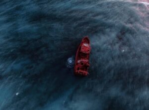 An aerial view of a rusted shipwreck adrift in dark blue waters, creating a stark contrast with its vibrant red hull.