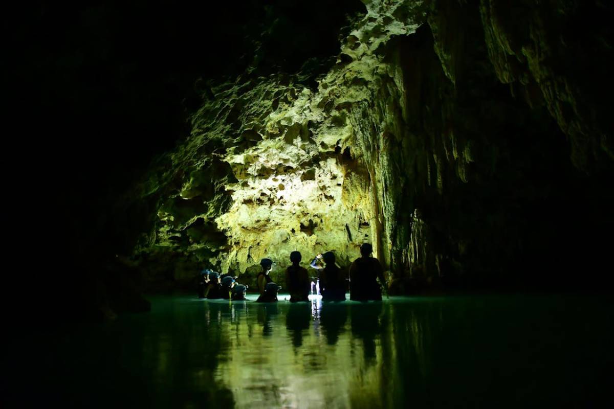 A group of silhouetted people explore a dark cave, illuminated by soft light reflecting off the water and rocky formations.