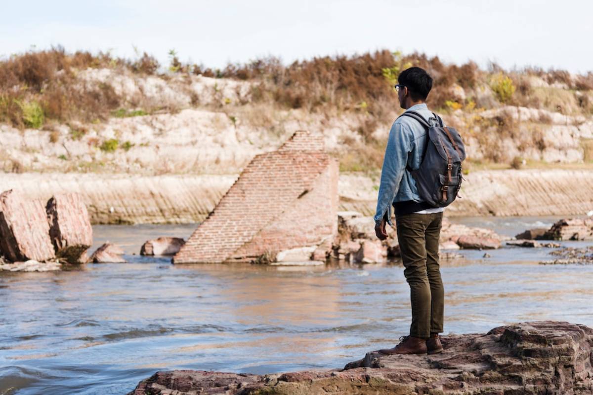 A person with a backpack stands on a rocky riverbank, gazing at a partially submerged brick structure in the water.