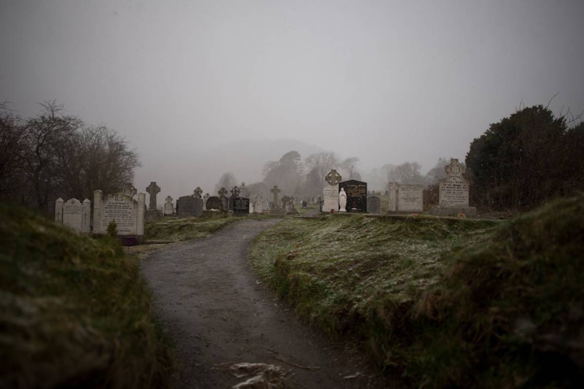 A misty graveyard path lined with weathered tombstones, surrounded by foggy hills and a soft layer of snow.