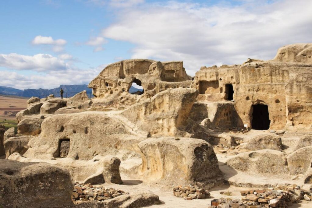 Ancient rock formations with caves and natural arches, set against a backdrop of mountains and blue sky.