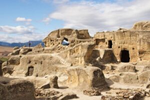 Ancient rock formations with caves and natural arches, set against a backdrop of mountains and blue sky.
