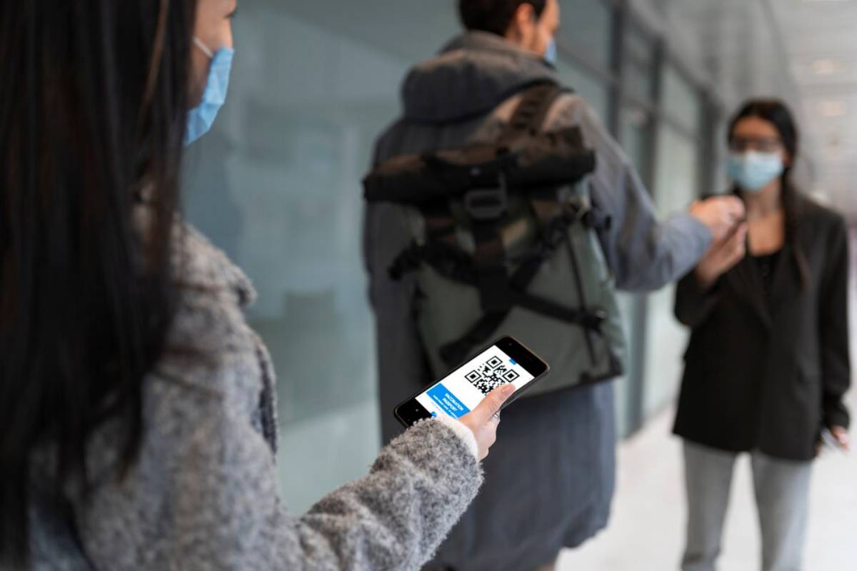 A person in a gray sweater holds a smartphone displaying a QR code, while another individual stands in the background.