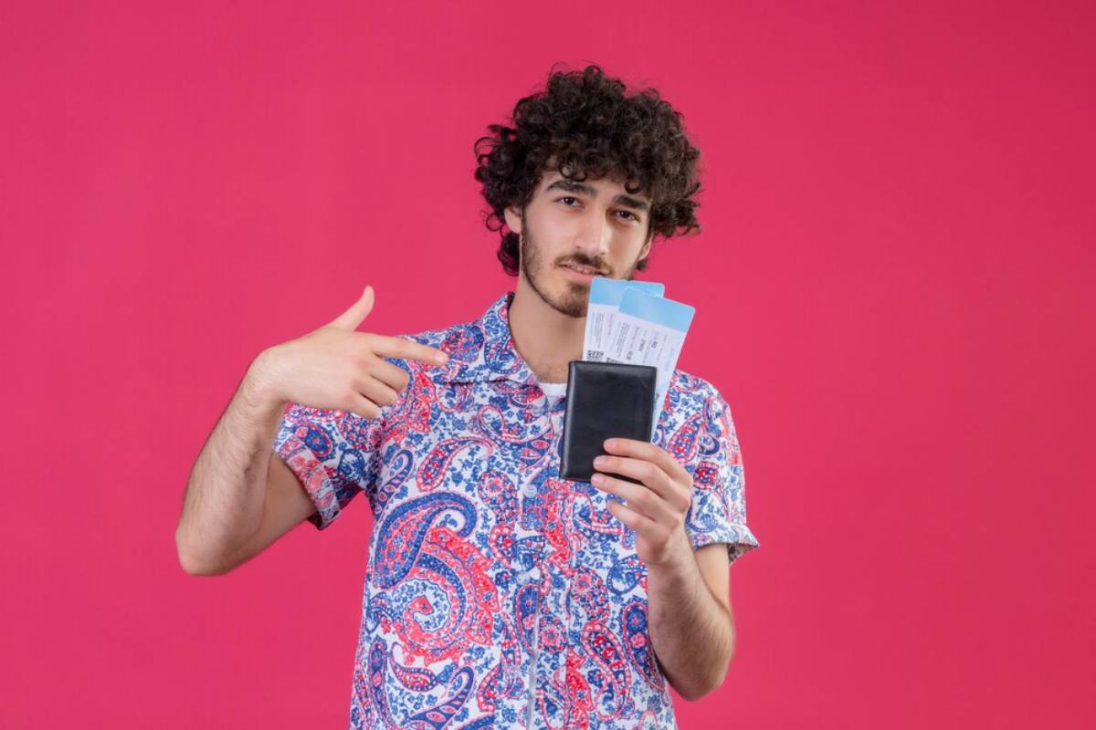 A person in a colorful paisley shirt holds a black wallet and flight tickets, smiling against a bright pink background.