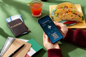 A hand holds a smartphone displaying a boarding pass, alongside a passport, tickets, tacos, and a cocktail on a green table.