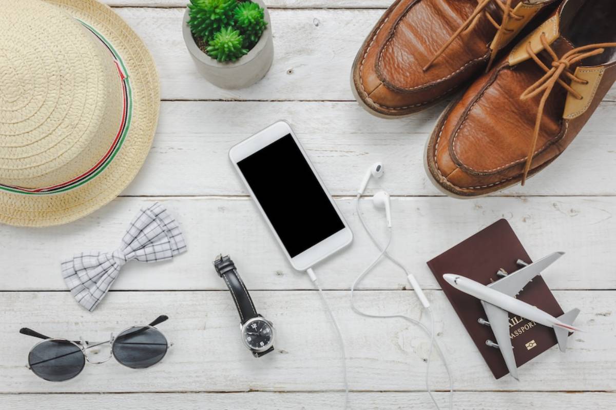Flat lay of travel essentials: straw hat, bow tie, sunglasses, smartphone with earphones, watch, brown shoes, passport, and airplane model.