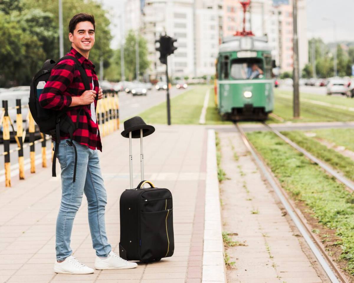 A young man in a red plaid shirt stands on a sidewalk next to a suitcase, waiting for a green tram on a busy street.