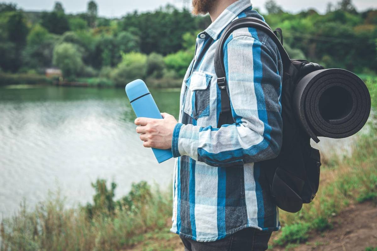 A man in a plaid shirt holds a blue thermos while standing near a lake, with a yoga mat rolled up on his backpack.