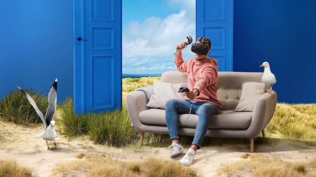 Person in a pink hoodie using a VR headset on a sofa, surrounded by a sandy landscape and a blue door, with seagulls nearby.