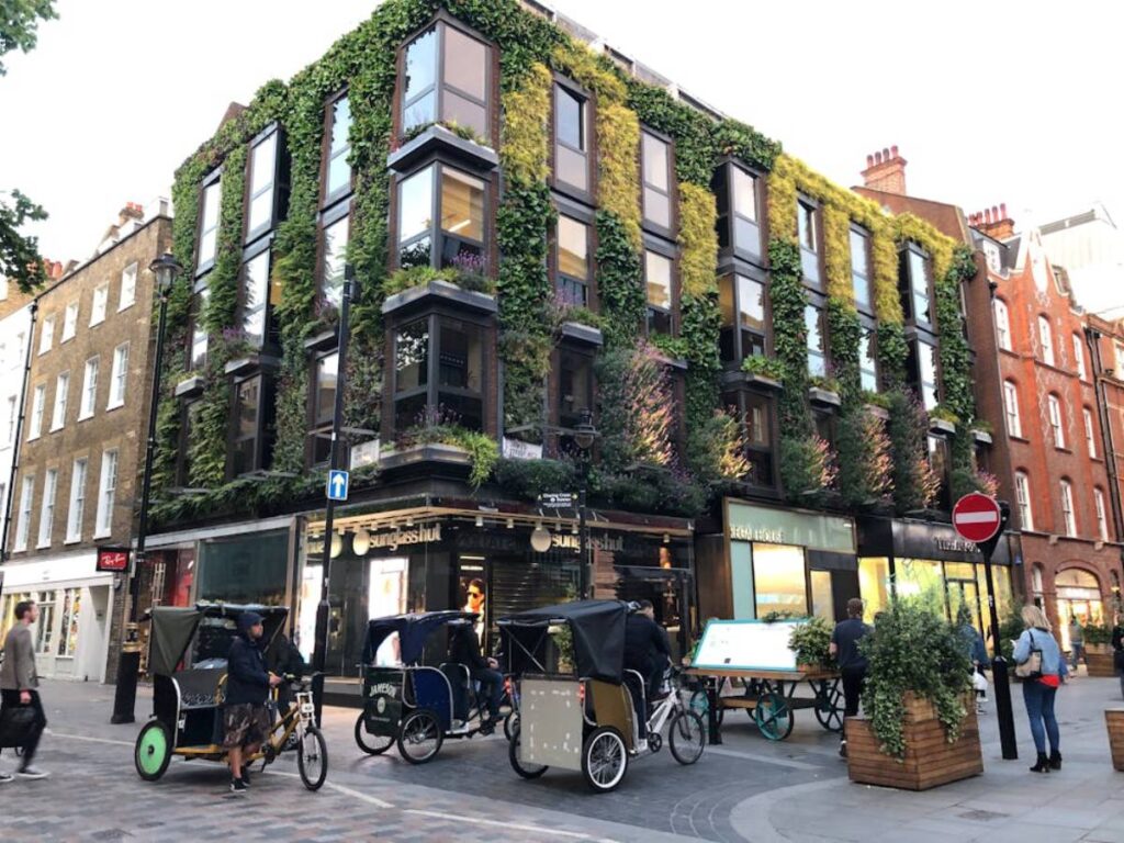 A modern building adorned with vibrant greenery, bustling cyclists in rickshaws, and pedestrians in an urban setting.