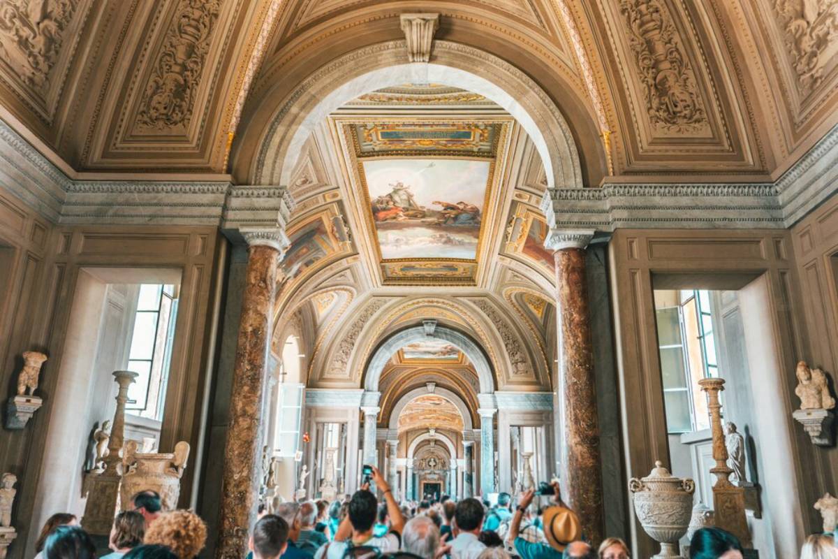 A bustling museum hallway adorned with ornate ceilings, marble columns, and sculptures, filled with visitors taking photos.