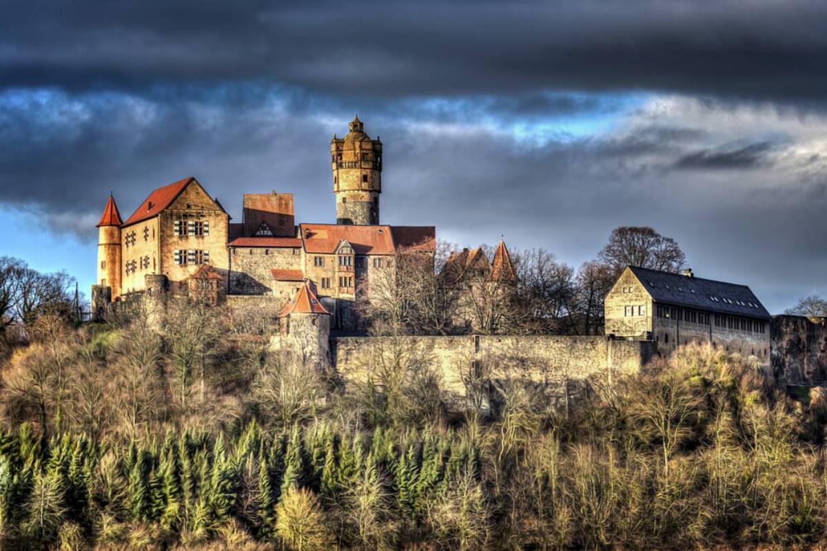 A historic castle perched on a hillside, surrounded by forested land and dramatic clouds in the sky.