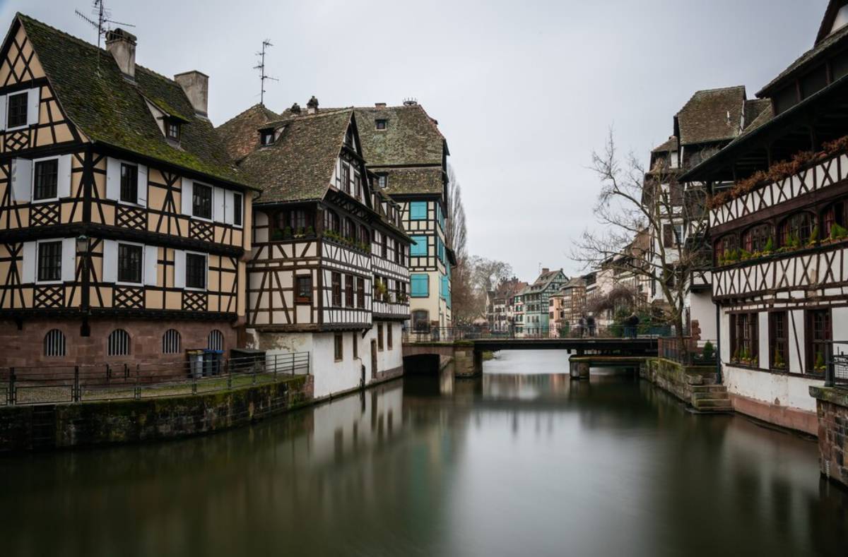 Half-timbered houses line a tranquil canal in Strasbourg, framed by a cloudy sky and reflections on the water's surface.