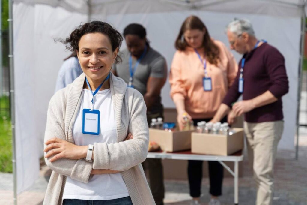 A group of volunteers works behind a table with supplies while one volunteer stands confidently in front, wearing a name tag.