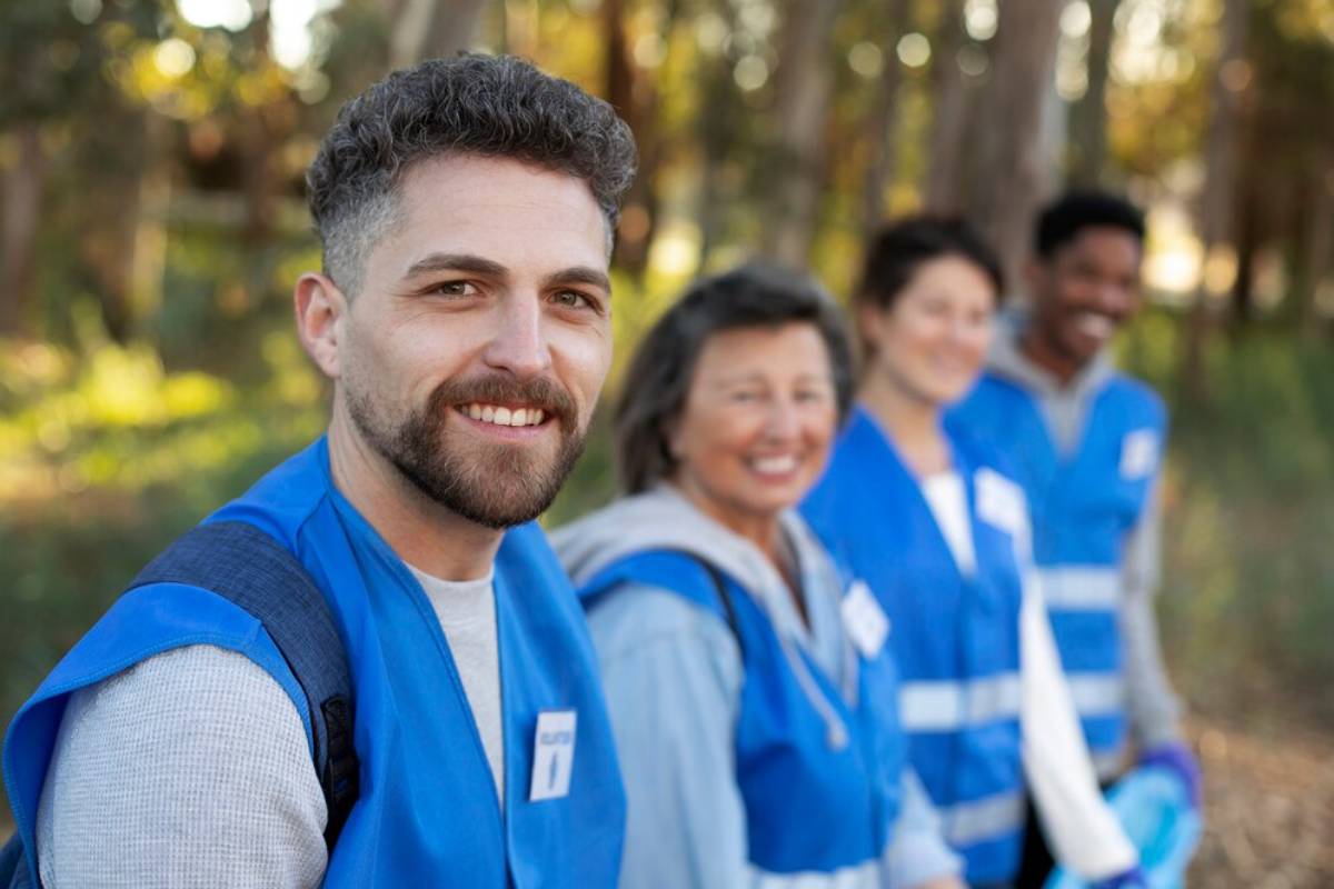 Four individuals wearing bright blue vests stand in a wooded area, promoting teamwork and community engagement in a sunny environment.