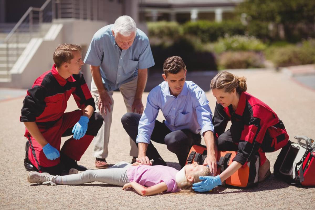 Emergency responders assist a young girl lying on the ground, surrounded by concerned individuals in a public space.
