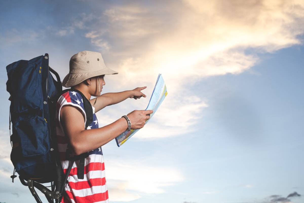 A person in a patriotic shirt and hat points at a map, standing against a beautiful sky at dusk, with a backpack slung over their shoulder.