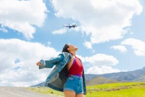 A woman with open arms stands in a field, gazing up at an airplane flying overhead against a bright blue sky and fluffy clouds.