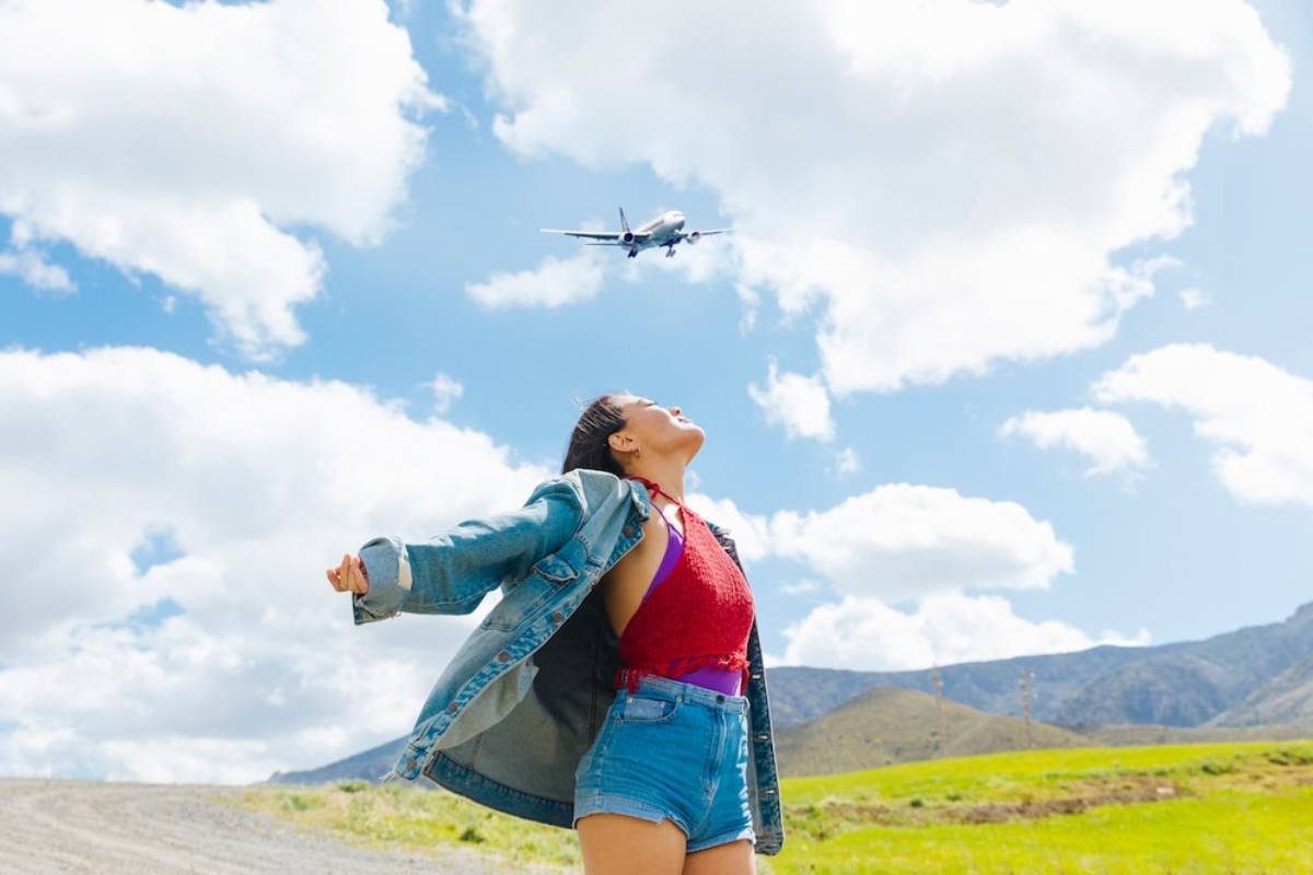 A woman with open arms stands in a field, gazing up at an airplane flying overhead against a bright blue sky and fluffy clouds.