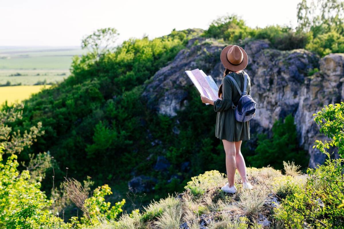 A young woman in a hat stands on a cliff, studying a map with green hills and a bright landscape in the background.