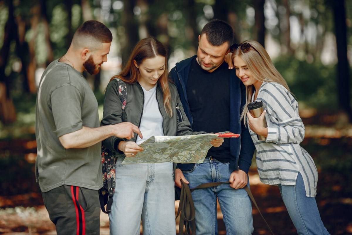 A group of four people studies a map together in a lush, green forest, surrounded by trees and dappled sunlight.