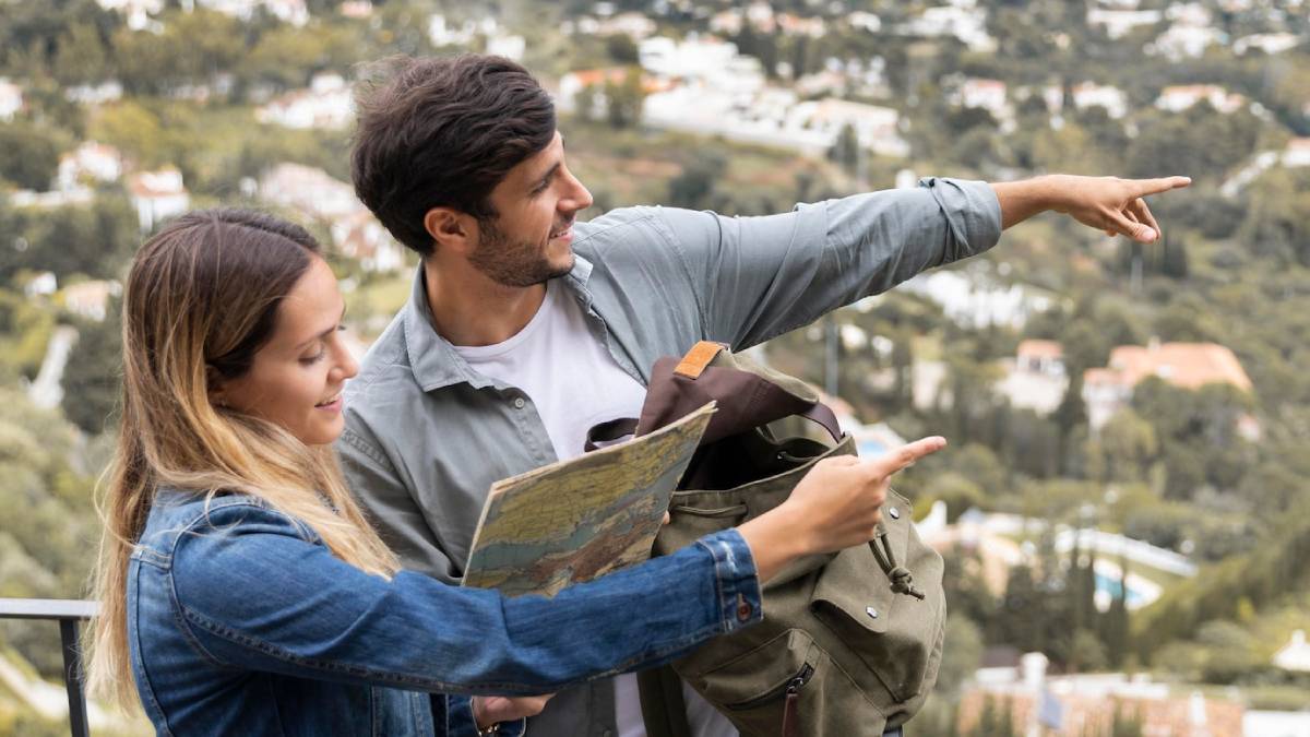 A couple stands outdoors, looking at a map while pointing out a location among scenic, lush greenery and distant houses.