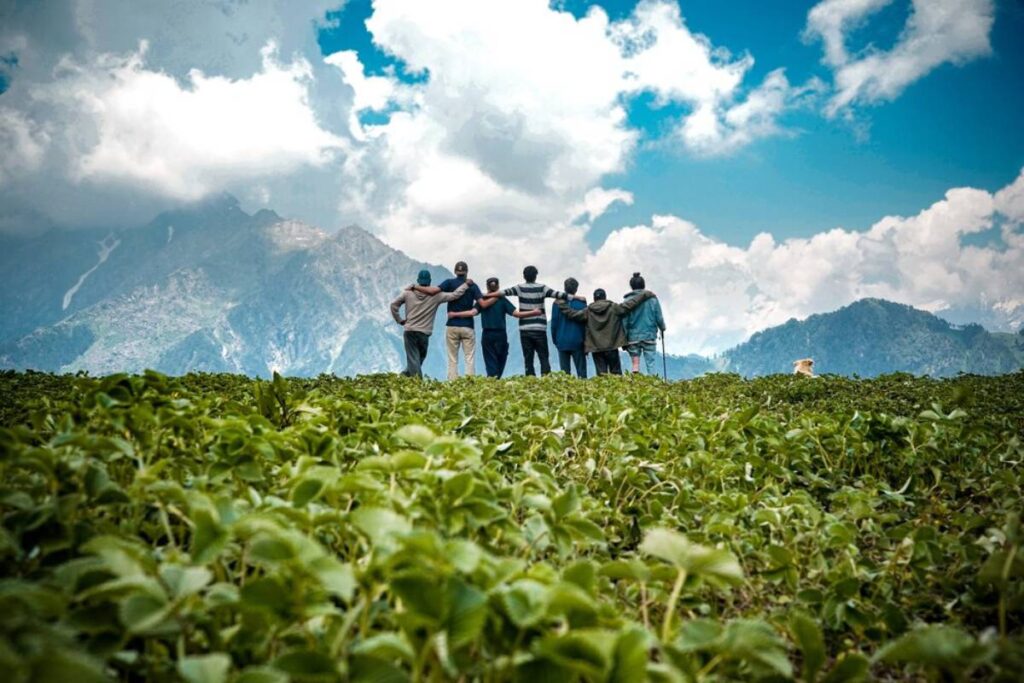A group of eight friends stands arm-in-arm on a lush green field, gazing at majestic mountains under a bright, cloudy sky.