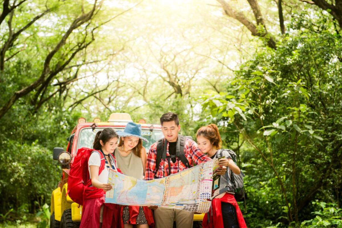 A group of four hikers studies a map surrounded by lush greenery, with a colorful off-road vehicle parked behind them.
