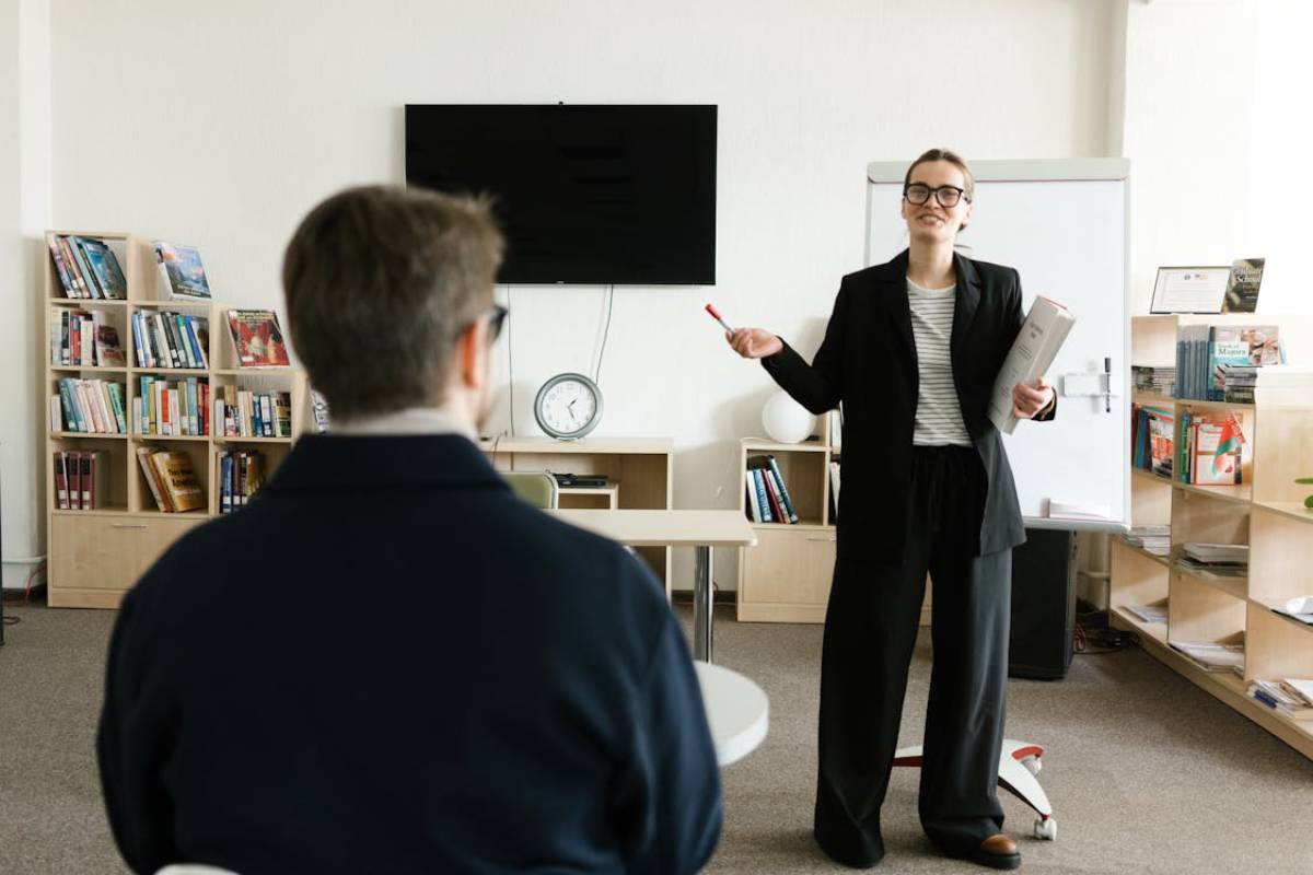A woman in a black suit stands in front of a classroom, gesturing with a pen and holding papers, while a person sits attentively.
