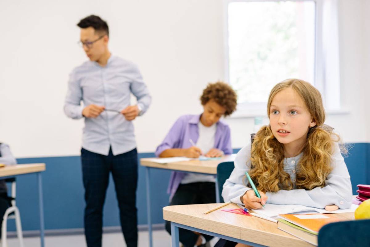 A classroom scene with students engaged in writing tasks, while a teacher stands, assisting in the background, surrounded by natural light.