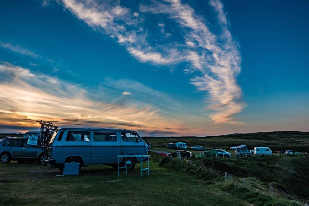 A vintage blue van parked on grassy terrain, with other vehicles and tents in the background under a colorful sunset sky.