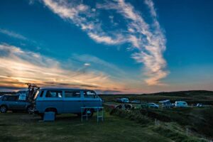 A vintage blue van parked on grassy terrain, with other vehicles and tents in the background under a colorful sunset sky.