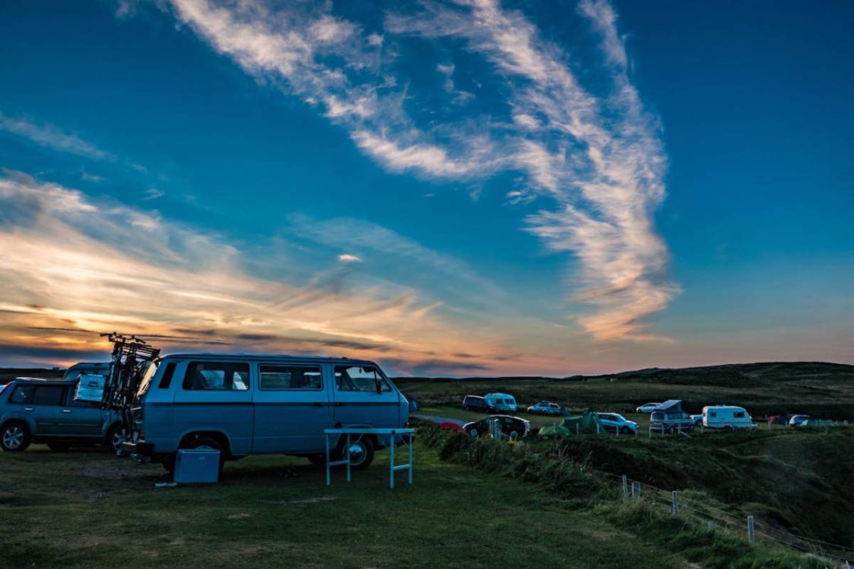 A vintage blue van parked on grassy terrain, with other vehicles and tents in the background under a colorful sunset sky.