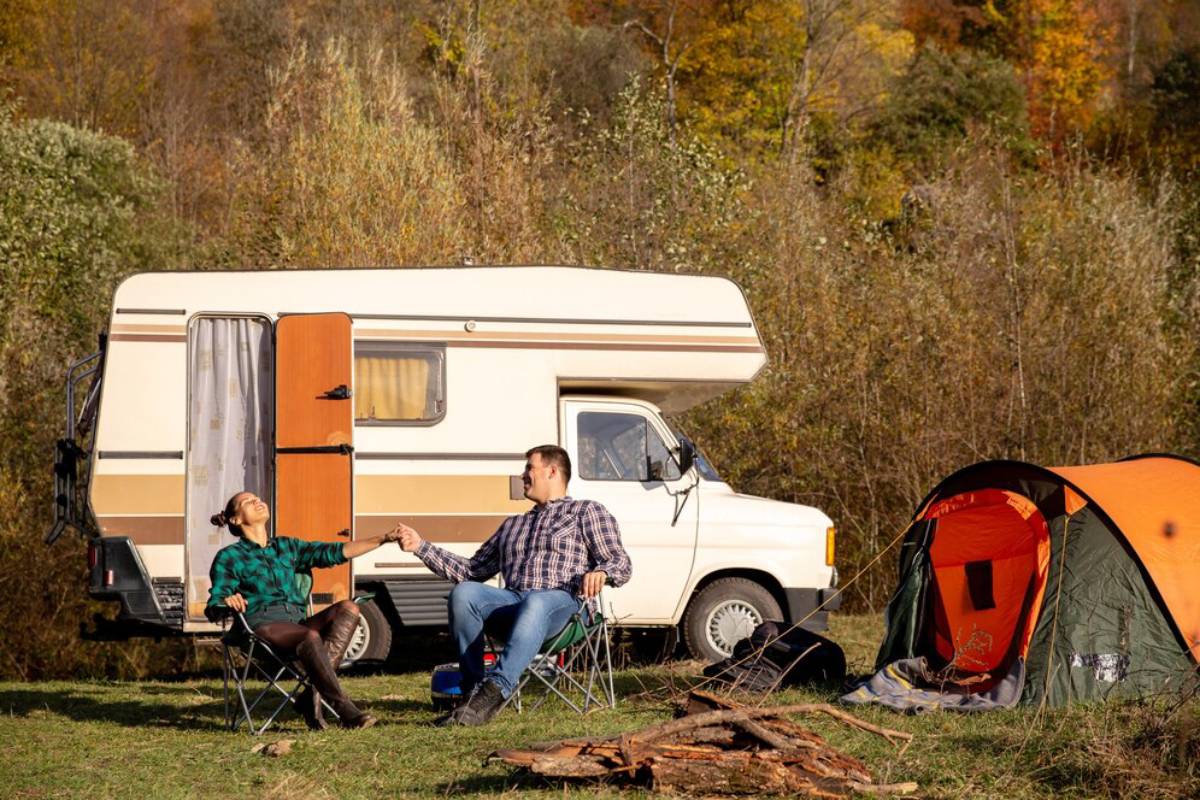 A couple sits by a campfire near a camping van and a tent, enjoying nature amidst autumn-colored trees.