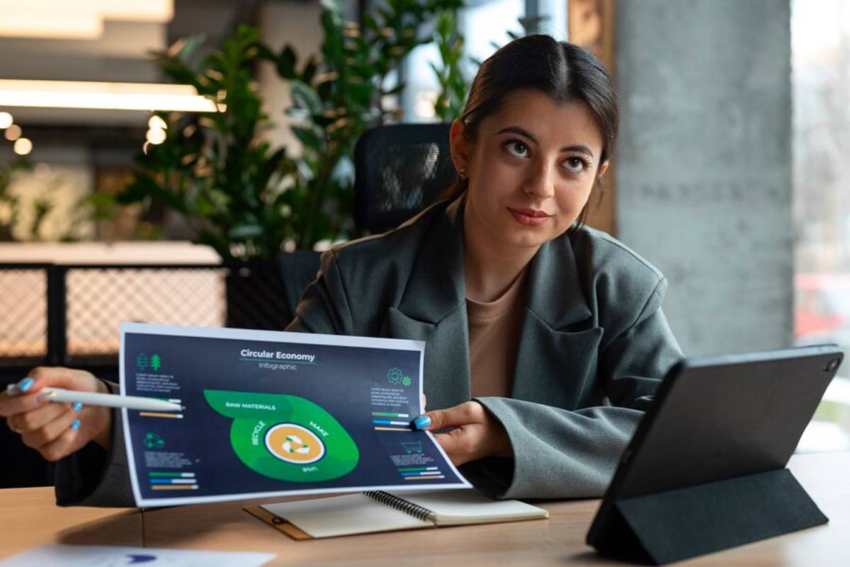 A person in a blazer holds an infographic on the circular economy, with a tablet and notebook on the table in a modern workspace.