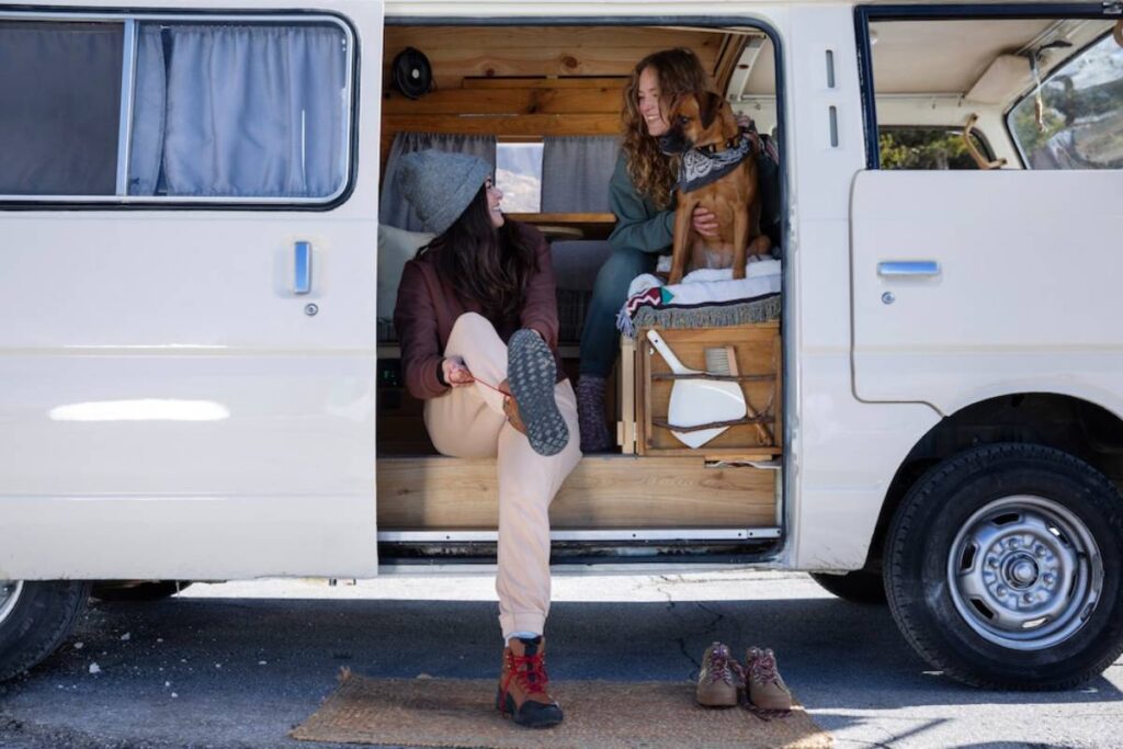 Two women relax in a camper van, one playfully adjusting her boot, while a dog sits on a blanket beside them. Outdoor adventure vibes.