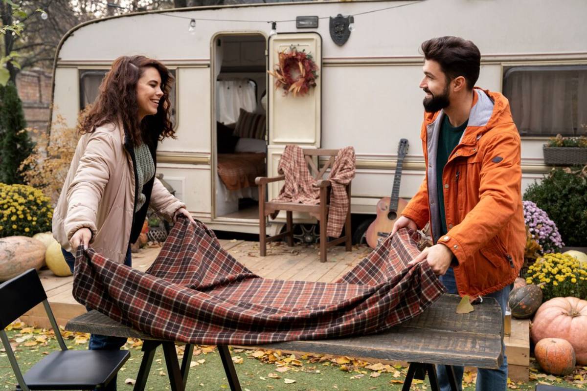 A couple prepares a plaid tablecloth on a rustic wooden table outside an inviting caravan surrounded by fall decorations.