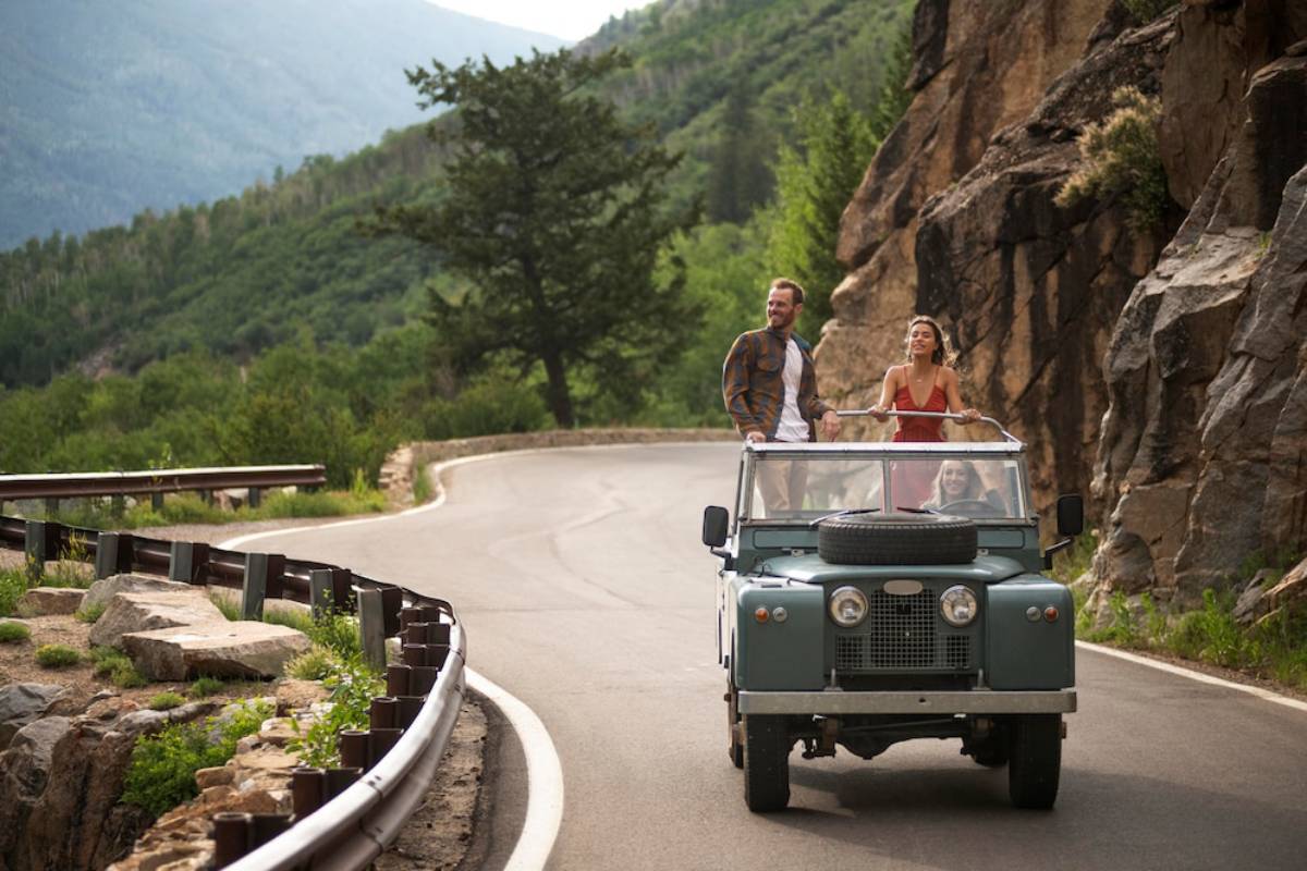 A vintage light blue convertible drives along a scenic mountain road, surrounded by lush greenery and rocky cliffs.