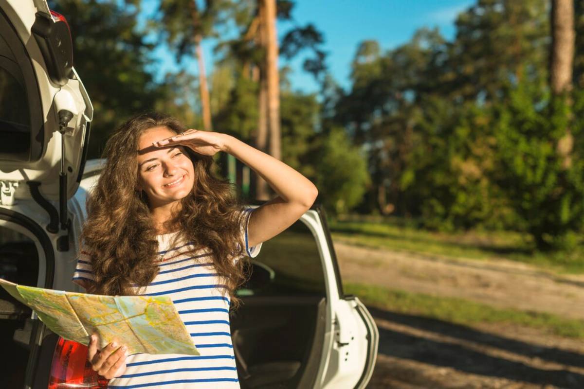 A woman stands next to a car in a forest, holding a map and looking thoughtful under a clear blue sky.