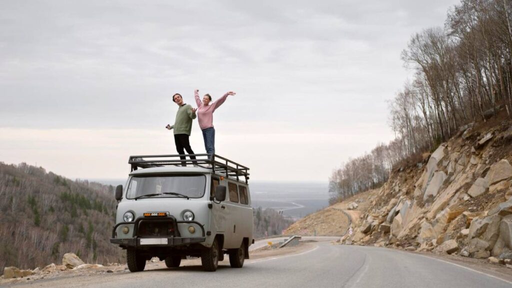 A couple joyfully poses on top of a vintage van parked by a winding road, surrounded by a scenic landscape of trees and hills.