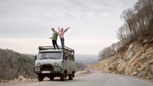 A couple joyfully poses on top of a vintage van parked by a winding road, surrounded by a scenic landscape of trees and hills.